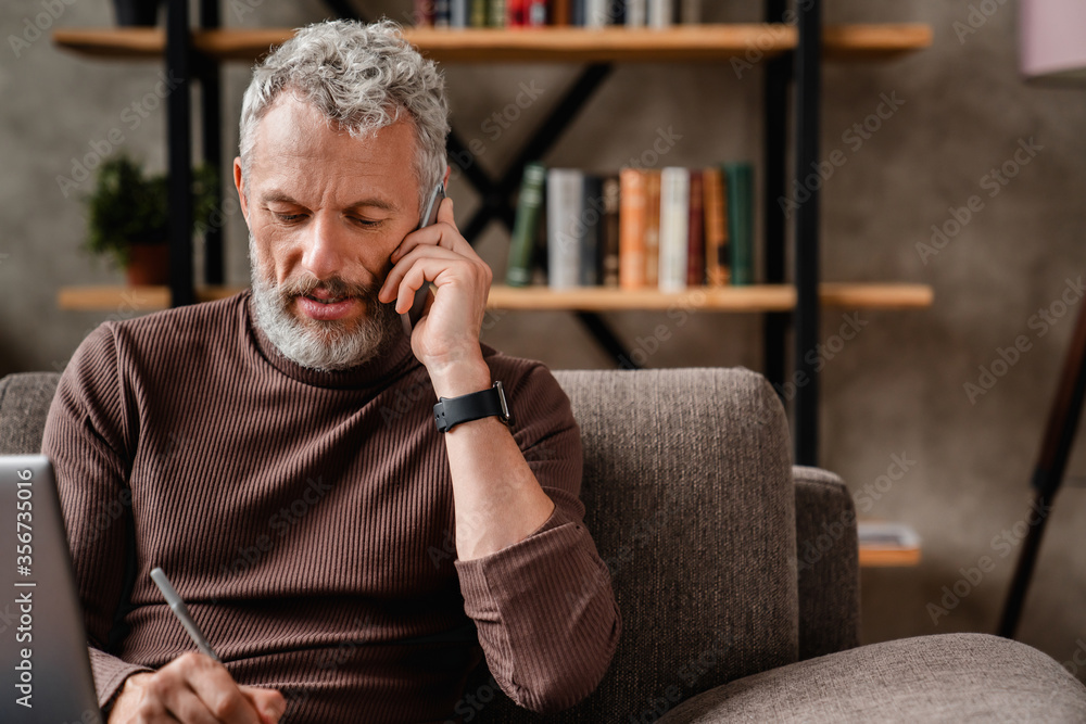 Wall mural Senior man in casual talking on mobile phone and writting notes while working at home using laptop