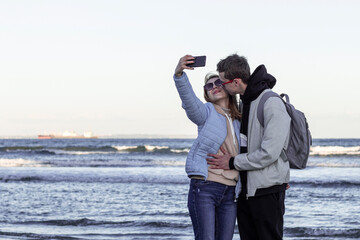 Obraz premium Young couple in love on sea shore taking selfie with mobile phone. Portrait of a romantic couple in love hugging and kissing by the sea. Tourists taking travel photos on their holidays in cold season.