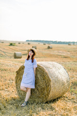 Naklejka na ściany i meble Countryside landscape of farm wheat field at summer sunset. Pretty young happy Caucasian woman in straw hat and blue striped dress posing near the hay bale and enjoying warm evening and vacations