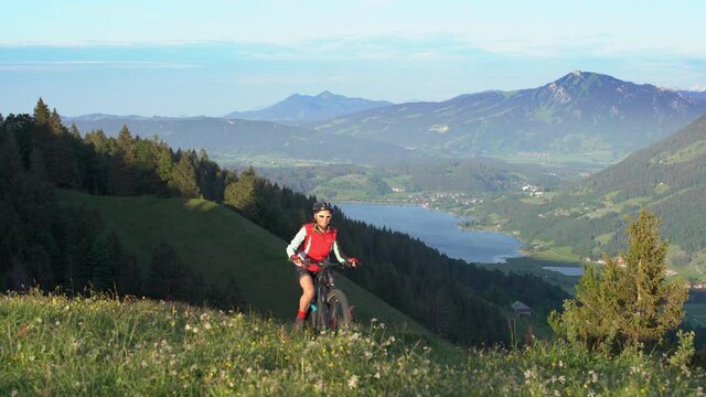 pretty senior woman riding her electric mountain bike on the mountains above Oberstaufen with lake Alpsee in the background, Allgau Alps, Bavaria Germany 