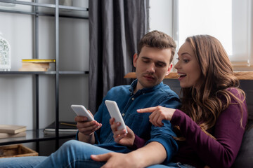 happy girl pointing with finger at smartphone near boyfriend in living room