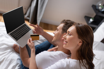 Obraz premium selective focus of cheerful woman holding credit card near man using laptop with blank screen in bedroom