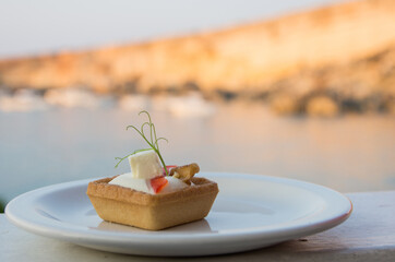 A single piece of finger food on a small white plate with a blurred rocks and sea in the background 