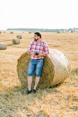 Naklejka na ściany i meble Handsome bearded brunette man, wearing checkered shirt and jeans leaning on a hay bale in goldern wheat field, posing, holding retro camera in hands, and enjoying warm sunset in summer day