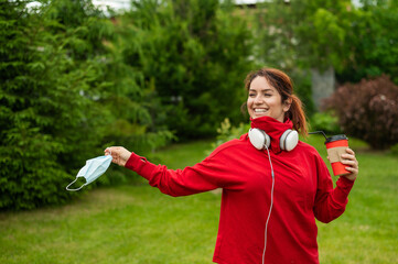 Red-haired woman in headphones and drinks coffee with a straw in the park and takes off a medical...