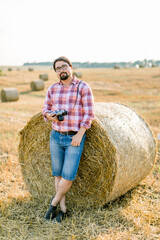 Naklejka na ściany i meble Full length portrait of handsome bearded young man in casual checkered shirt and jeans, standing outdoor in summer field, leaning on hay bale, and holding in hands retro camera