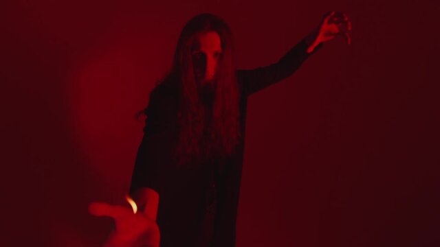 A young magician with long hair shows a hot hand to the camera. A flame of fire on the arm of an illusionist with long hair on a red background