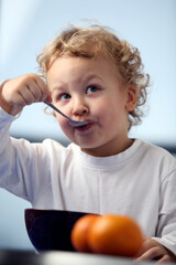 Portrait of the little funny, cheerful boy having a meal. Cute child in a white shirt with a big spoon having breakfast, lunch or dinner. 