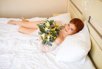 Obraz na płótnie Canvas The beautiful red-haired girl in a transparent peignoir lies on the bed with a bouquet in her hands. Boudoir morning of the bride. Top view.