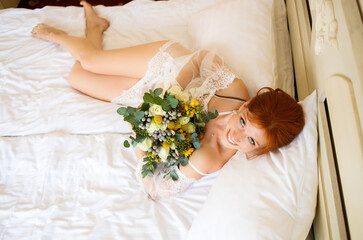 Obraz na płótnie Canvas The beautiful red-haired woman in a transparent peignoir lies on the bed with a bouquet in her hands. Boudoir morning of the bride. Top view.