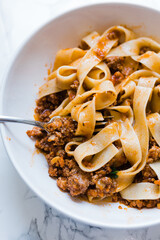 A closeup of a bowl of pappardelle noodles covered in bolognese sauce, a type of meat sauce from Italy. This delicious Italian cuisine dish is served in a white bowl with a fork.