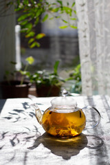 Glass transparent teapot with herbal tea on a background of a window with flowers.