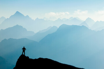 Man reaching summit after climbing and hiking enjoying freedom and looking towards mountains silhouettes panorama during sunrise.