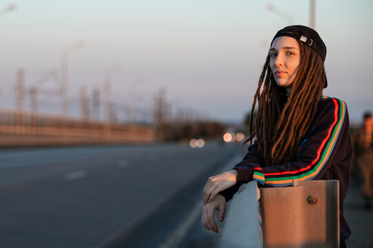 girl in a sports sweatshirt with dreadlocks in a cap looks out for a car on the road. hitch-hiking. stand by