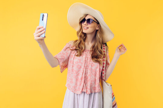 smiling young woman in a summer hat and sunglasses takes a selfie on a mobile phone on an yellow background