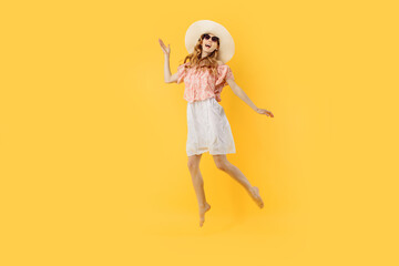 An excited happy girl in a summer hat and sunglasses jumps in the air with her fists raised, looking at the camera isolated on a yellow background