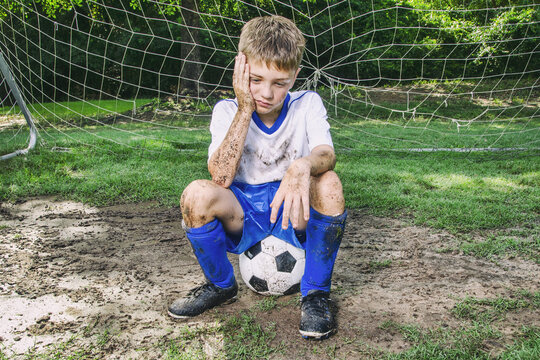 Little muddy soccer boy sitting unhappy in front of goal