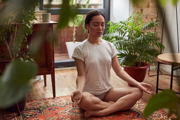 Pleasant female in underclothes meditating indoors surrounded by pot plants