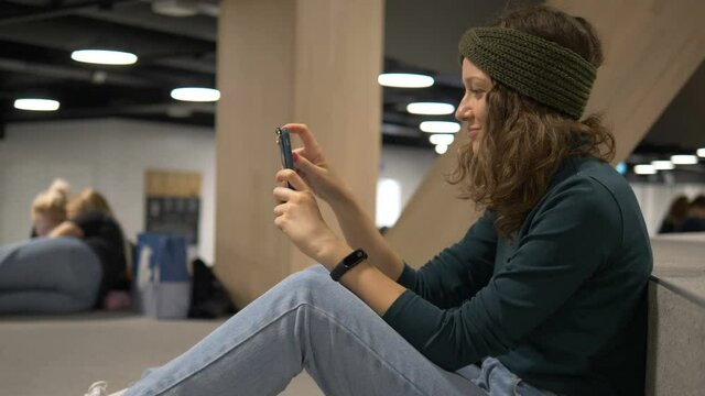 smiling modern woman uses smartphone with electronic clock, student sits on stairs in modern library in Helsinki