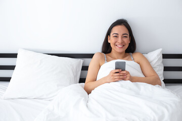 Obraz na płótnie Canvas Smiling young female relaxing with smartphone in bedroom