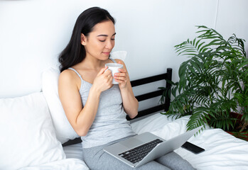 Calm young woman with laptop sipping tea in bedroom