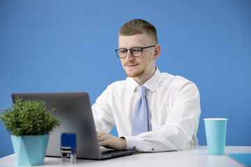 Portrait of a young caucasian male office worker with glasses and a white shirt working at a laptop. Modern stylish workplace. Isolated on a blue background.