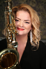 Obraz premium Portrait of a charming blonde with a saxophone in her hands. Beautiful girl in a black evening dress. Young woman with her hair and bright red lips. Face close up. Saxophonist lady