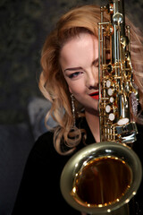 Obraz premium Portrait of a charming blonde with a saxophone in her hands. Beautiful girl in a black evening dress. Young woman with her hair and bright red lips. Face close up. Saxophonist lady