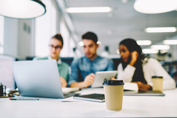 Craft paper mug with coffee to go standing on wooden table on blurred background with working group silhouette