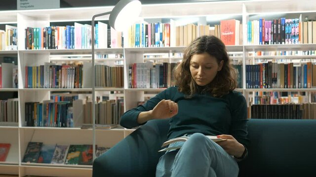 young beautiful woman sitting on a sofa and reading a book in a cozy public library on background of bookshelves