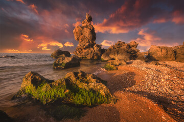 Fantastic sunset on the picturesque rocky sea coast. Beautiful seascape with sharp cliffs, calm water, golden beach and amazing colors on the sky.