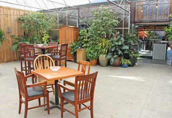Practising social distancing during Covid-19 pandemic. The view on the empty restaurant wooden tables and chairs on the sunny patio. Flower and green plant pots in the background.