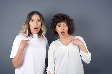 Surprised beautiful female student being in stupor shocked, has astonished expression pointing at herself with finger saying: Who me? Isolated over gray background.