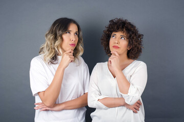 Shot of contemplative thoughtful Young woman keeps hand under chin, looks thoughtfully upwards, dressed in casual clothes, poses over white background with free space for your text