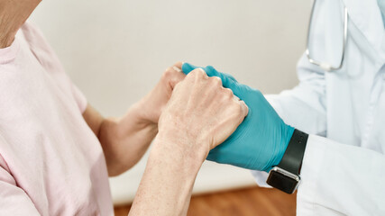 Cropped shot of a doctor in blue sterile gloves holding hands of female senior patient. Main focus on hands. Close up. Healthcare