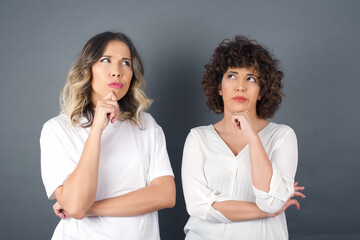 Isolated portrait of stylish young European woman with hand under chin and looking sideways with doubtful and skeptical expression, suspect and doubt. Standing indoors over background.