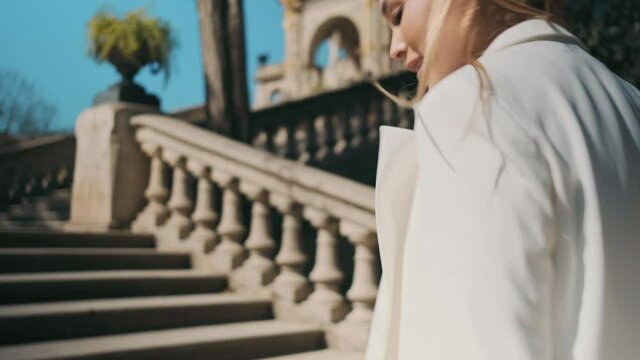 Tracking shot of young gorgeous woman in classic white suit confidently walking up the stairs in old city park and showing follow me gesture on camera