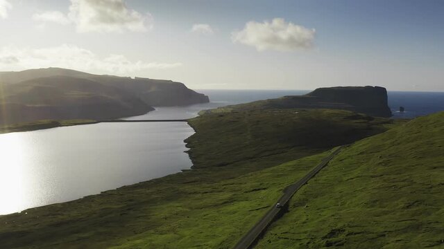 Car driving along coastal road view on wild green island at sunset.Cinematic rear view approaching.Faroe Island road along beautiful coast. Faroe island, Denmark. Forward aerial flight