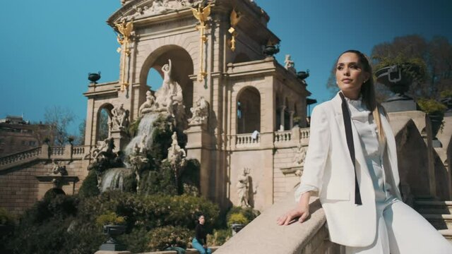 Tracking shot of young posh woman in white suit confidently posing on stairs in park with old beautiful architecture