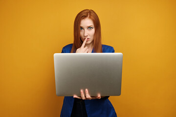 Naklejka premium A red-haired girl in a blue jacket stands on a yellow background with a laptop in hand and putting finger to lips