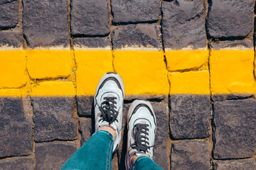 women's feet in white sneakers stand on the pavement one foot stepped on a bright yellow line,...
