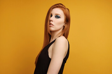 Red-haired girl in a black top and with blue eye makeup is standing on a yellow background with hair covering half her face