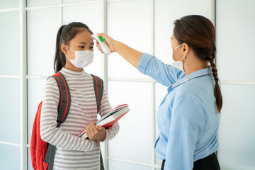 Asian woman teacher using thermometer temperature screening student for fever against the spread of COVID-19 while student coming back to school, New normal and Education concept.