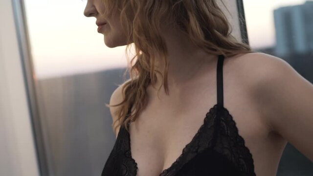 Close up of woman in a black dress with a deep neckline on her chest. Action. Caucasian seductive female wearing black negligee with curly hair standing by the window