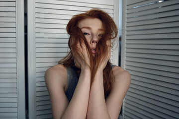 A red-haired girl in a t-shirt and pants sits against a white screen and looks at the camera, covering her face with hair.