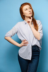 Red-haired girl in a striped shirt stands on a blue background and thoughtfully raising a finger to...