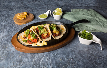 Tacos al pastor accompanied by pineapple, guacamole and spicy sauce served in an iron pan.