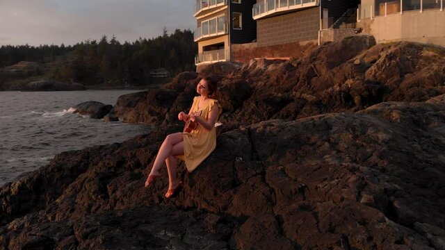 Young attractive brunette woman playing a ukulele by the ocean at sunset. 4k 24fps.