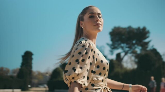 Tracking around shot of young posh woman in polka dot dress confidently looking in camera walking along park