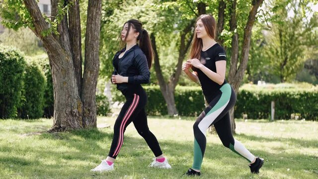 Two young ladies are training together at the park. The friends are doing lunges to strengthen their legs and booty.